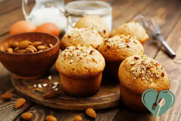 Freshly baked almond flour muffins topped with chopped nuts, surrounded by whole almonds, eggs, and milk on a wooden table.