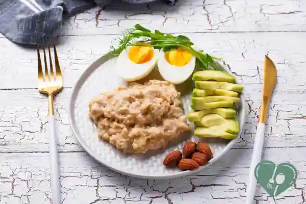 A diabetic-friendly breakfast plate with steel-cut oats, sliced avocado, a boiled egg, almonds, and fresh greens, arranged on a white rustic wooden surface.