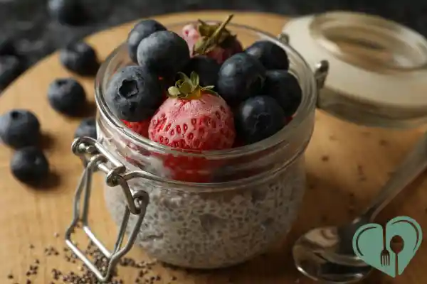 A glass jar filled with chia pudding, topped with fresh blueberries and a whole strawberry, placed on a wooden board with a spoon nearby.
