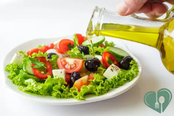Hand pouring olive oil over a fresh Mediterranean salad with feta, olives, and greens