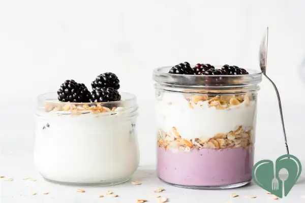 Two glass jars filled with Greek yogurt layered with berries and oats, topped with blackberries and served with a spoon.