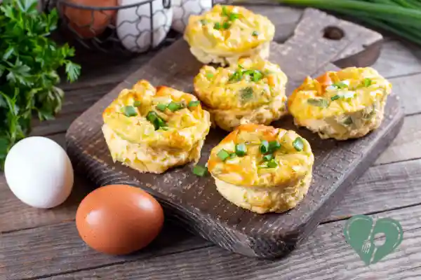 Baked egg muffins with chopped green onions on a wooden board, surrounded by raw eggs and fresh herbs.