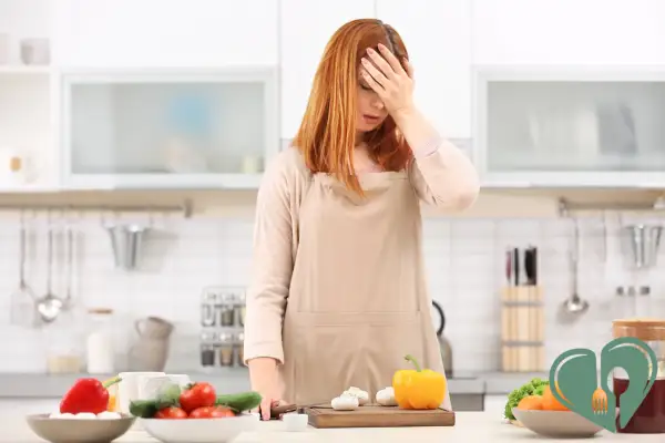 A tired woman standing in the kitchen with her hand on her forehead, surrounded by vegetables and prep ingredients on the counter.