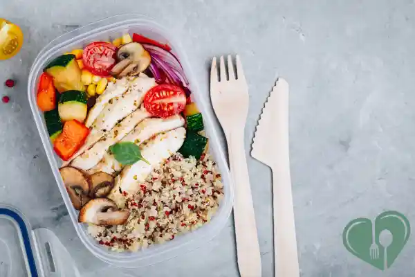 Meal prep container with grilled chicken, quinoa, and colorful vegetables beside wooden utensils.
