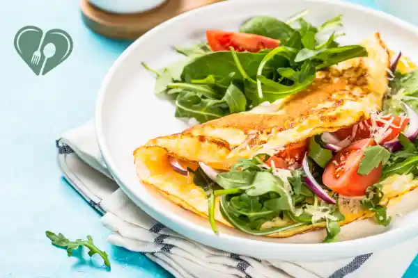 Golden omelet folded over fresh arugula, cherry tomatoes, and red onion on a white plate, served with greens on a blue background.