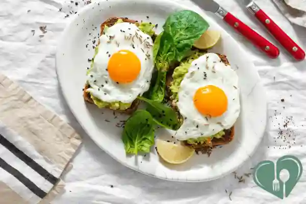 Two slices of whole grain toast topped with avocado, fried eggs, and fresh spinach leaves on a white plate.