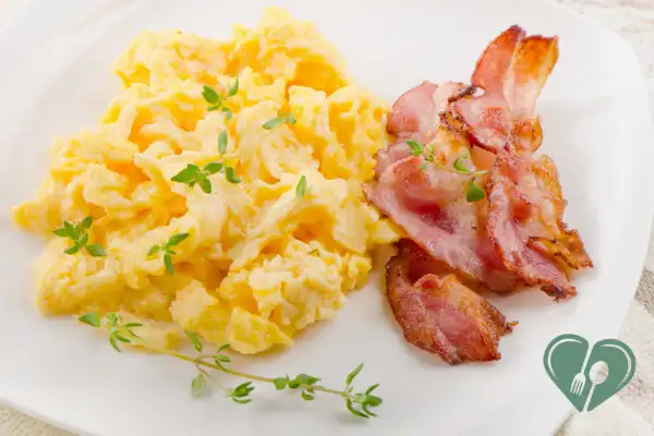 Scrambled eggs with crispy bacon strips, garnished with fresh thyme on a white plate.