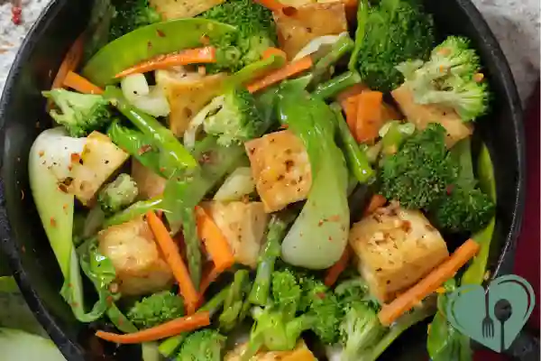 Tofu cubes stir-fried with broccoli, snap peas, carrots, and bok choy in a cast iron skillet, sprinkled with chili flakes.