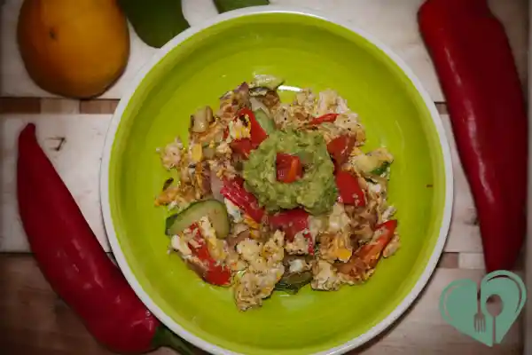 A green bowl filled with tofu scramble, sautéed peppers, zucchini, and onions, topped with a scoop of guacamole and surrounded by red bell peppers and a mango.