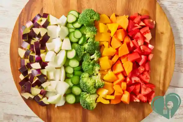 Freshly chopped vegetables – eggplant, onion, cucumber, broccoli, yellow pepper, and tomato – arranged on a wooden board.